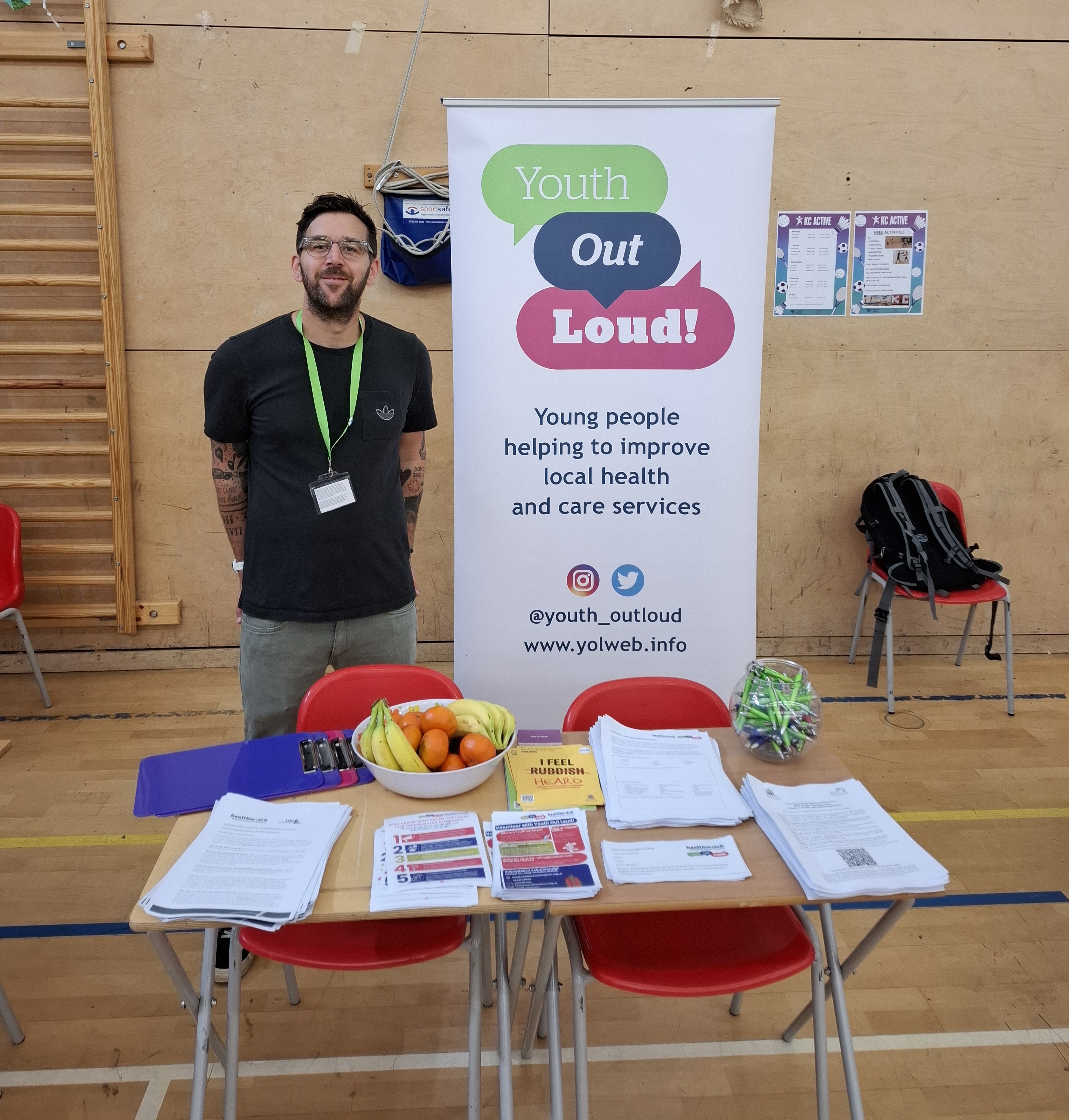 A photo of a man stood behind and information stall with a banner for 'Youth Out Loud!' and an an information stall with fresh fruit. The stall appears to be in a school gymnasium.