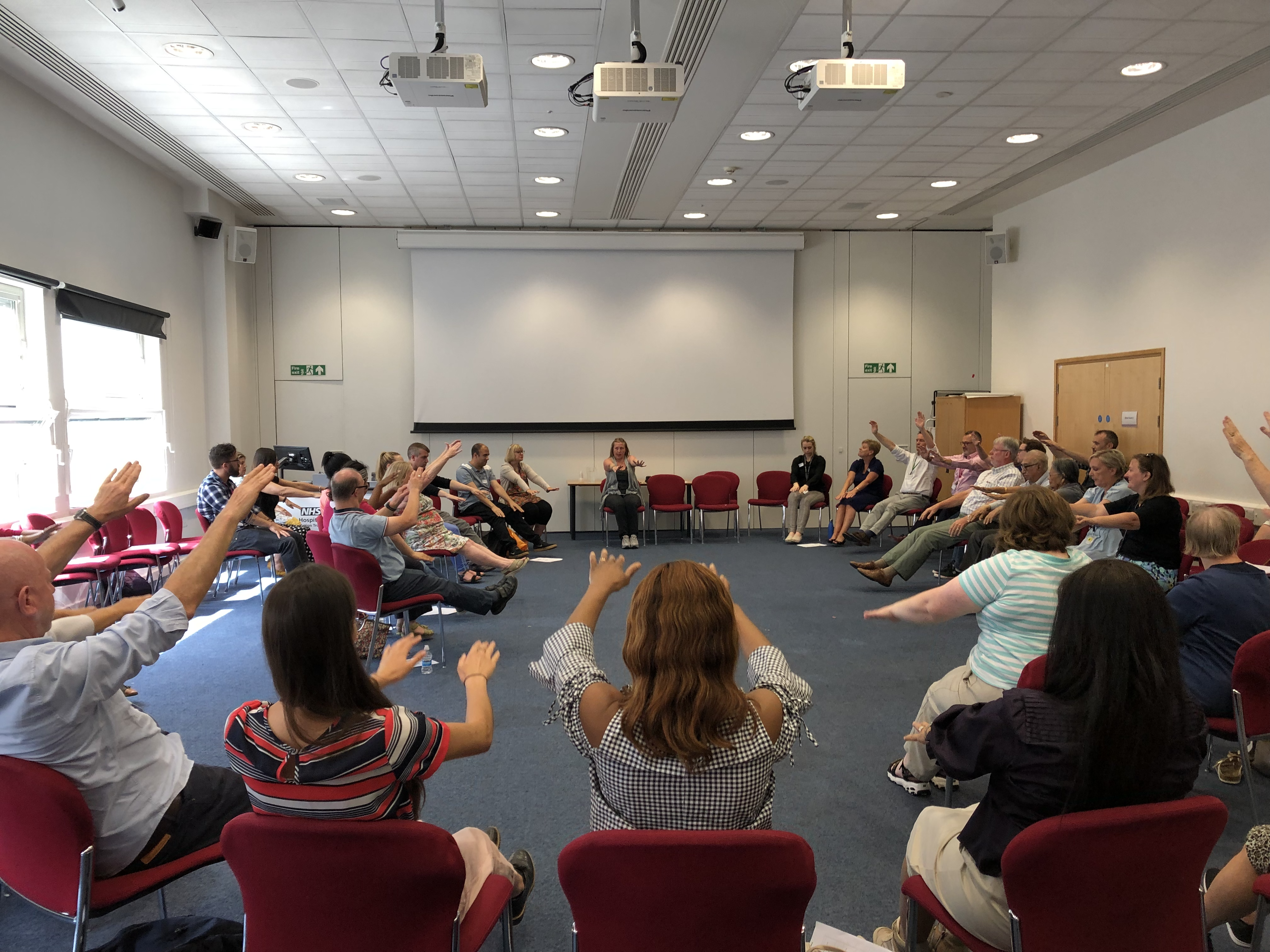 A photo of a group of people sat in a circle on chairs in a large room, they all have their arms outstretched in a group activity.