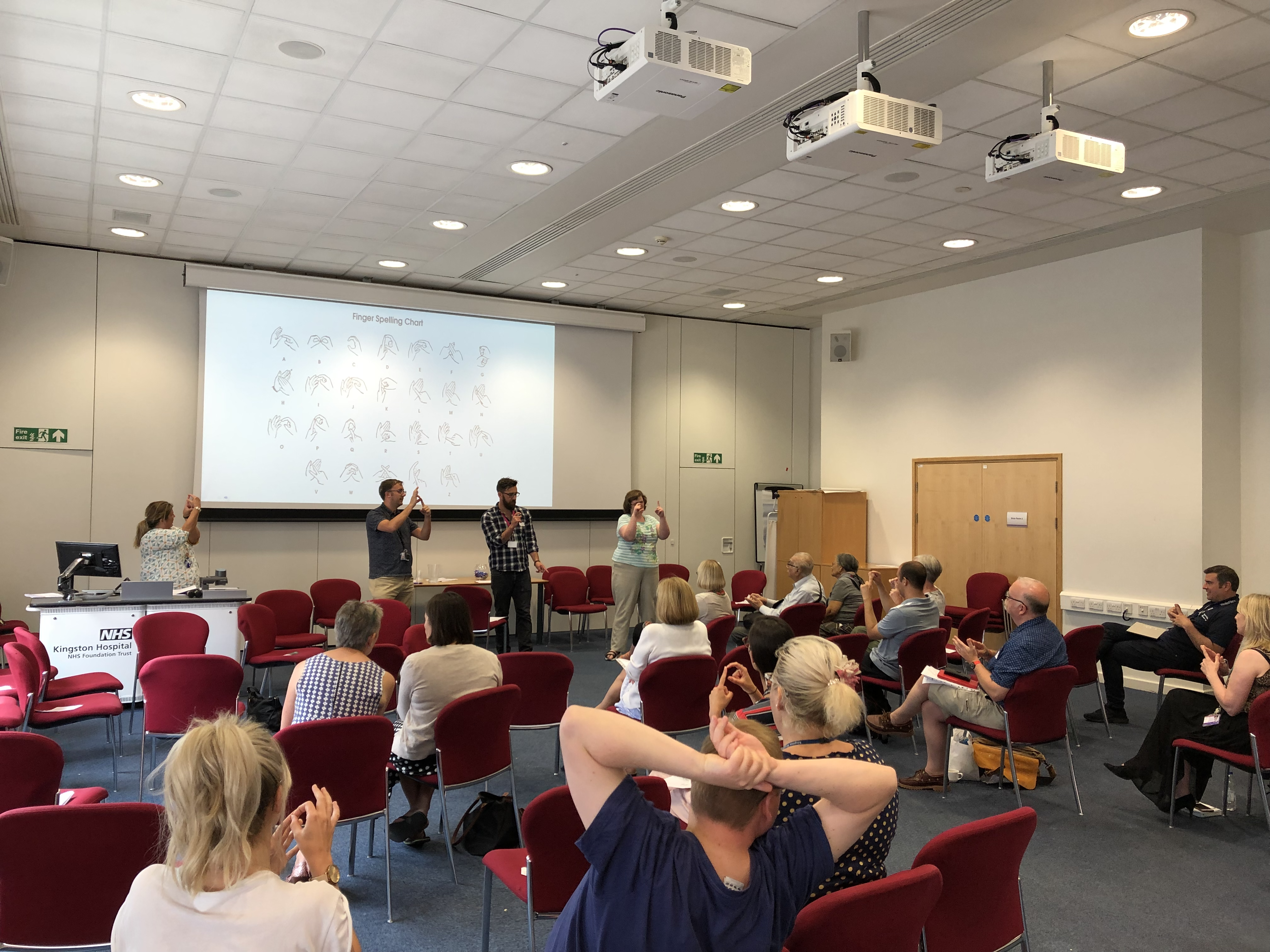 A photo of a group of people in large room, they are sat on chairs facing 3 people at the front of the room who are finger spelling. Behind them the finger spelling alphabet is projected on to the wall.