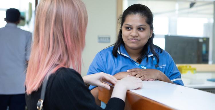 A photo of a woman at reception desk speaking to a worker in medical scrubs