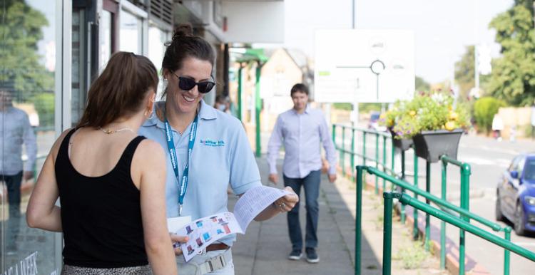 A photo of a two women speaking outside a public building. One is wearing a Healthwatch branded polo and lanyard while holding an information leaflet.