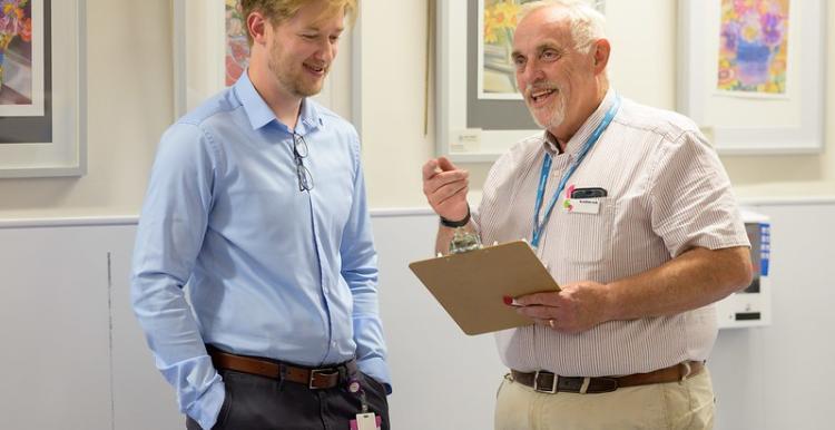 A photo of two people talking to each other, one is holding a clipboard and pointing