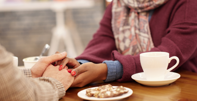 A photo of two women sat in a cafe, one holds the other's hands in hers over the table in support.