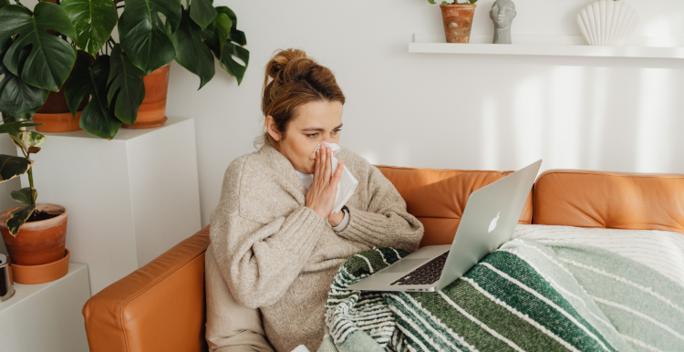 A photo of a woman blowing her nose, she is wrapped up in a blanket and has a laptop on her lap.