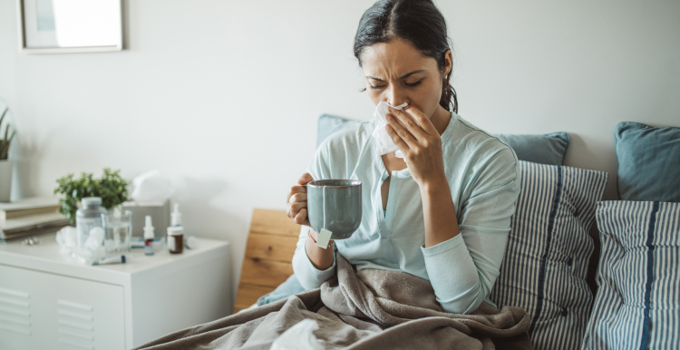 A photo of a woman sat upright in bed blowing her nose. She is holding a cup of tea and there are medicines on her bedside table. 