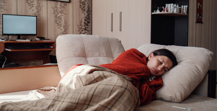 A photo of a woman looking unwell as she huddles under blankets. 
