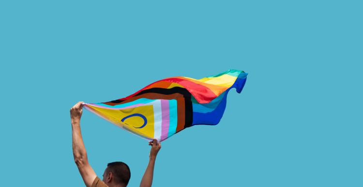 A photo of a person holding a billowing Pride flag against a backdrop of blue sky.