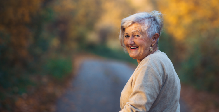 A photo of an older woman turning to smile. She appears to be on a path surrounded by woodland. 