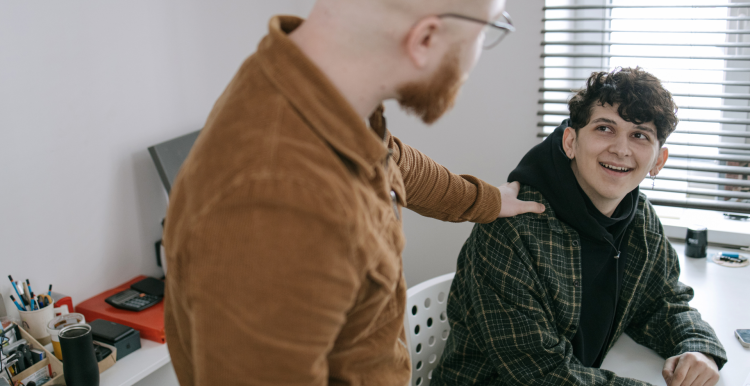A photo of a supportive adult putting their hand on the shoulder of a younger person who is looking up at them and smiling.