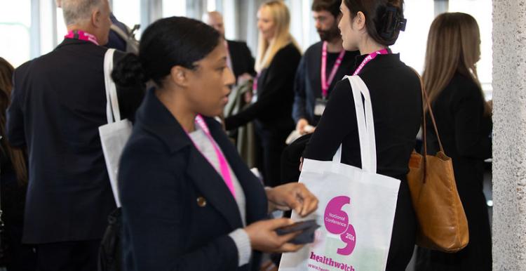 A photo of a group of people smartly dressed appearing to network. They are wearing white tote bags with bright pink healthwatch branding.