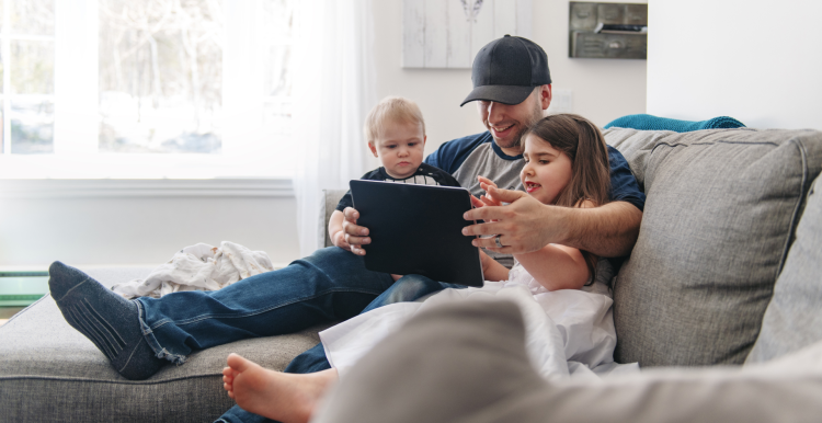 A photo of a dad with two children. They are sat on a sofa, he has his arms around a toddler and an older child as they watch a tablet together.