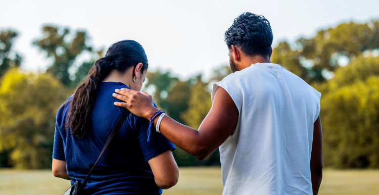 A photo of two people in a green space. Their backs are to the camera and one has their hand on the others shoulder in a comforting gesture. Both their heads are bowed slightly. 