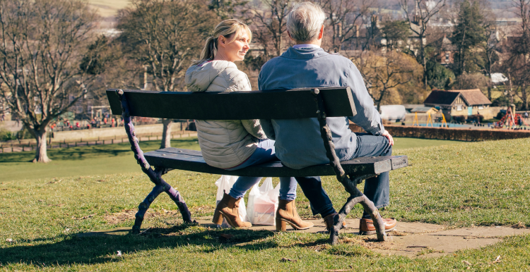 A photo of two people sat on a bench in a green space. One has her face turned toward the other gesturing that she is listening.