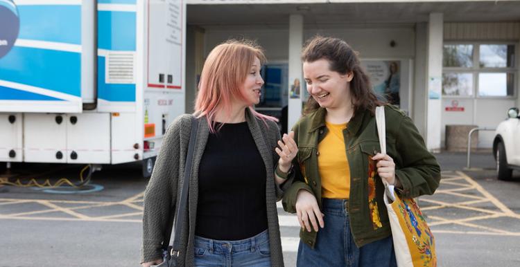 A photo of two women walking away from a hospital, they are smiling and laughing as they talk to each other. 