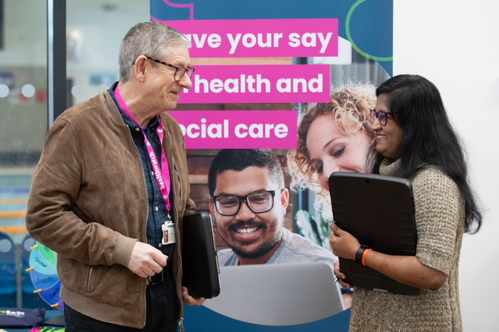 A photo of two people stood in front of a Healthwatch branded banner that reads 'Have your say on health and social care'