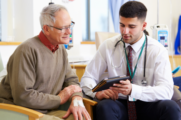 A photo of an older man talking to a doctor, the doctor is using a digital tablet to share information with the patient.