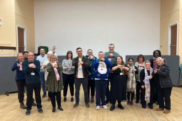 A photo of a large group of people, they stand facing the camera with the arms outstretched putting one hand over the other. Many are wearing lanyards.