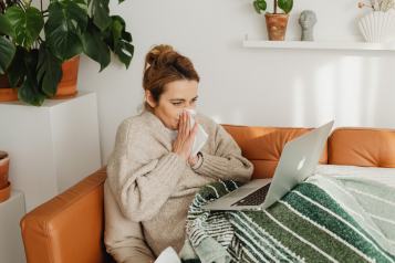 A photo of a woman blowing her nose, she is wrapped up in a blanket and has a laptop on her lap.