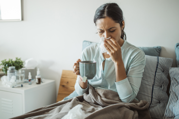 A photo of a woman sat upright in bed blowing her nose. She is holding a cup of tea and there are medicines on her bedside table. 