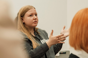 A photo of a woman talking, she is using her hands to gesture and we see the back of another woman's head facing the same way. 