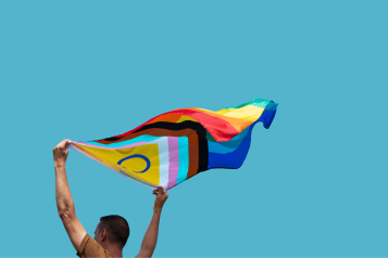 A photo of a person holding a billowing Pride flag against a backdrop of blue sky.