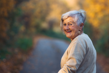 A photo of an older woman turning to smile. She appears to be on a path surrounded by woodland. 