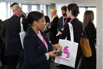 A photo of a group of people smartly dressed appearing to network. They are wearing white tote bags with bright pink healthwatch branding.