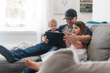 A photo of a dad with two children. They are sat on a sofa, he has his arms around a toddler and an older child as they watch a tablet together.