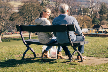 A photo of two people sat on a bench in a green space. One has her face turned toward the other gesturing that she is listening.