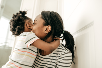 A photo of a woman with a young child. The child is hugging her and the woman's face is turned away from us.