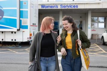 A photo of two women walking away from a hospital, they are smiling and laughing as they talk to each other. 