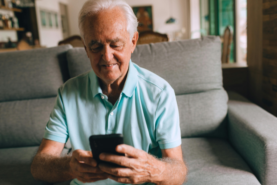 A photo of an older man using his phone in a living space, his expression is unclear