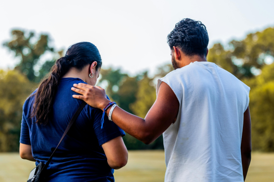 A photo of two people in a green space. Their backs are to the camera and one has their hand on the others shoulder in a comforting gesture. Both their heads are bowed slightly. 