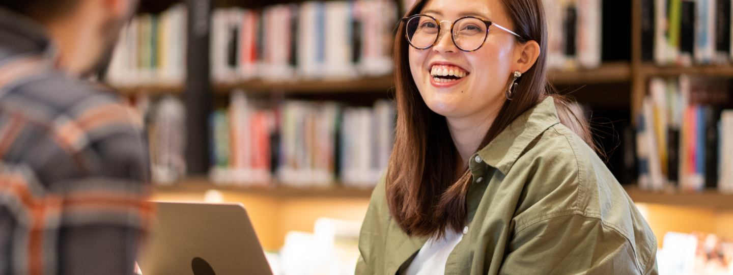 A photo of a young woman in a library or bookshop smiling as she looks at someone who is just out of frame