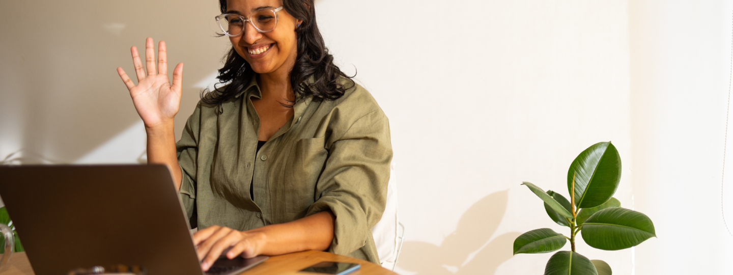 A photo of a woman looking waving at her laptop, she is in a room lit by natural light and there is a houseplant on the floor next to her