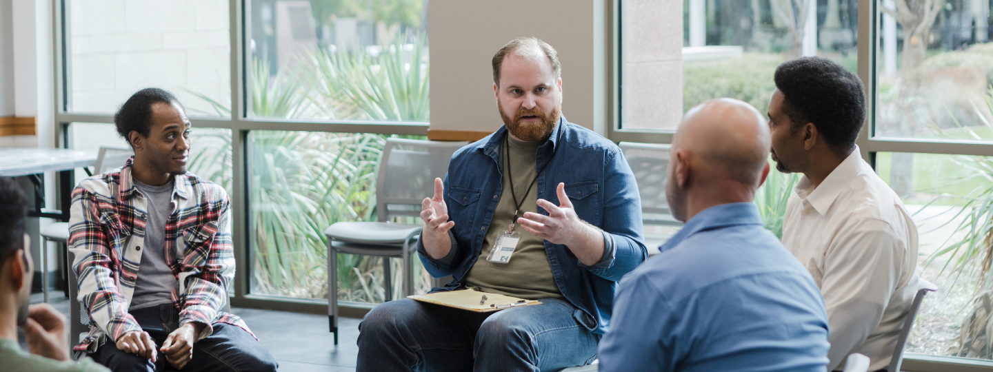 A photo of men in a support group. A man with a lanyard sits in the centre of the image and leads the discussion