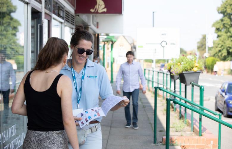 A photo of a two women speaking outside a public building. One is wearing a Healthwatch branded polo and lanyard while holding an information leaflet.