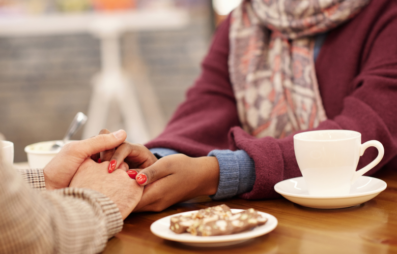 A photo of two women sat in a cafe, one holds the other's hands in hers over the table in support.