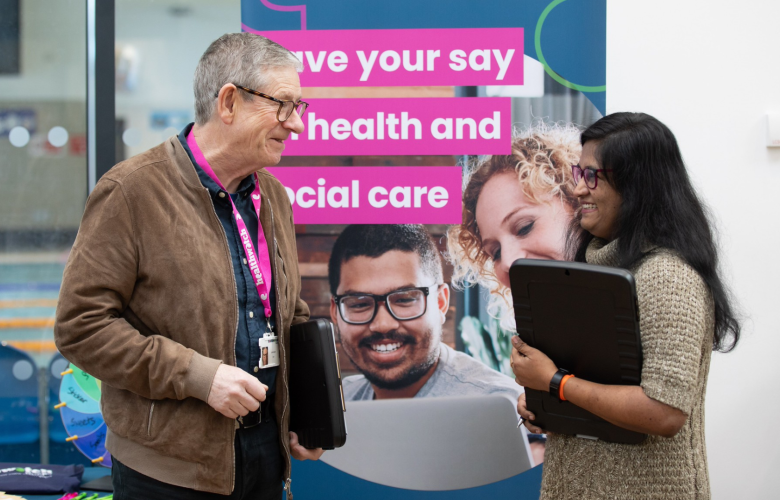 A photo of two people stood in front of a Healthwatch branded banner that reads 'Have your say on health and social care'