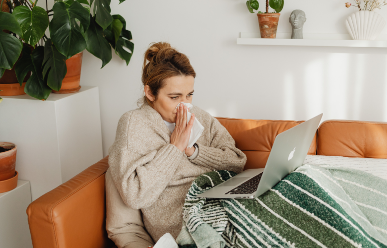 A photo of a woman blowing her nose, she is wrapped up in a blanket and has a laptop on her lap.