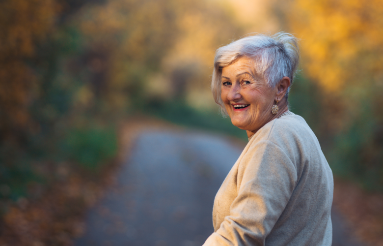 A photo of an older woman turning to smile. She appears to be on a path surrounded by woodland. 