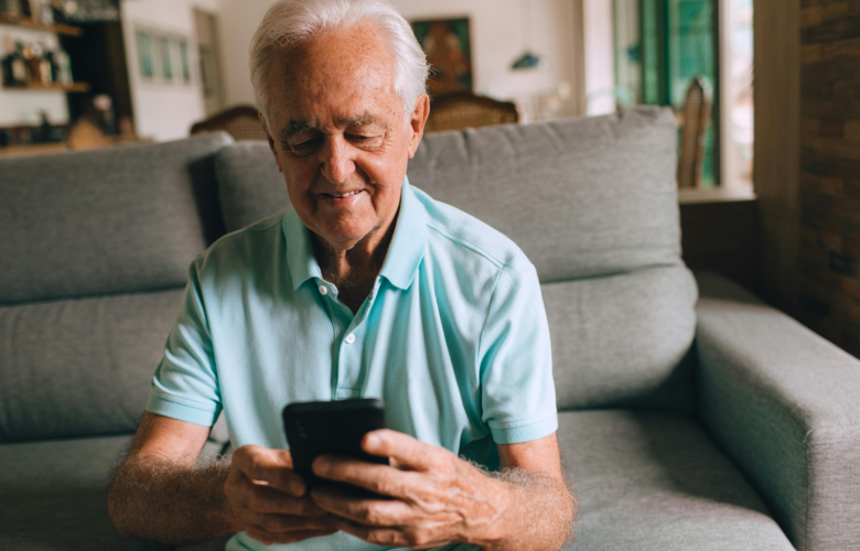 A photo of an older man using his phone in a living space, his expression is unclear