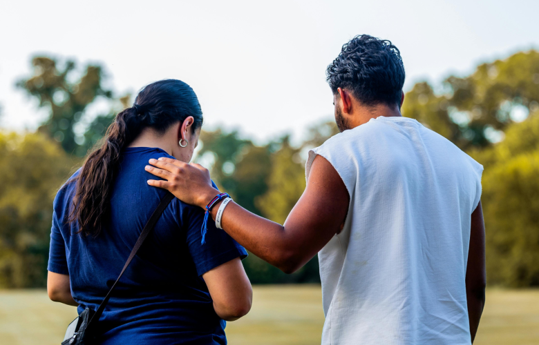 A photo of two people in a green space. Their backs are to the camera and one has their hand on the others shoulder in a comforting gesture. Both their heads are bowed slightly. 