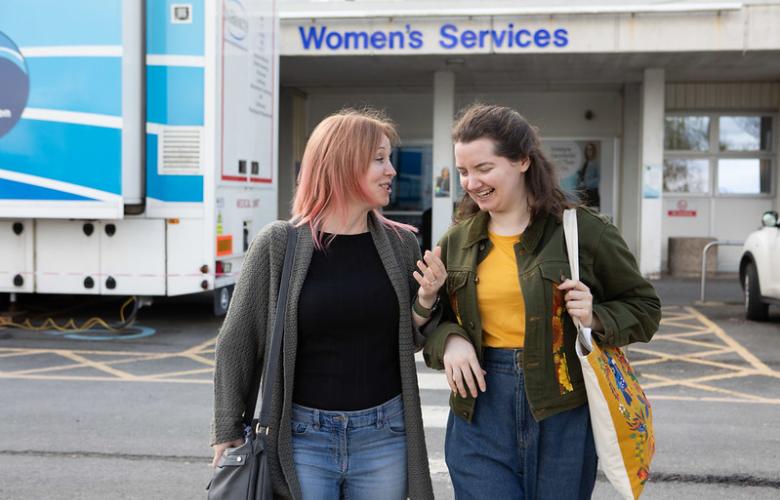 A photo of two women walking away from a hospital, they are smiling and laughing as they talk to each other. 